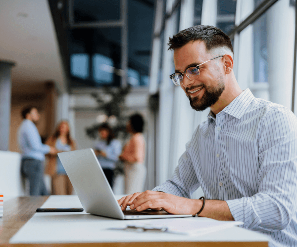 Άτομο κάθεται σε γραφείο σε σύγχρονο επαγγελματικό περιβάλλον και εργάζεται σε laptop, ενώ στο φόντο φαίνονται συνάδελφοι που συζητούν όρθιοι.