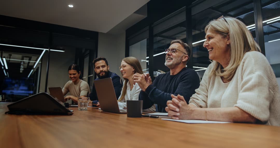 Ομάδα επαγγελματιών σε αίθουσα συσκέψεων συμμετέχει σε συνάντηση γύρω από τραπέζι, με laptops και σημειώσεις μπροστά τους.