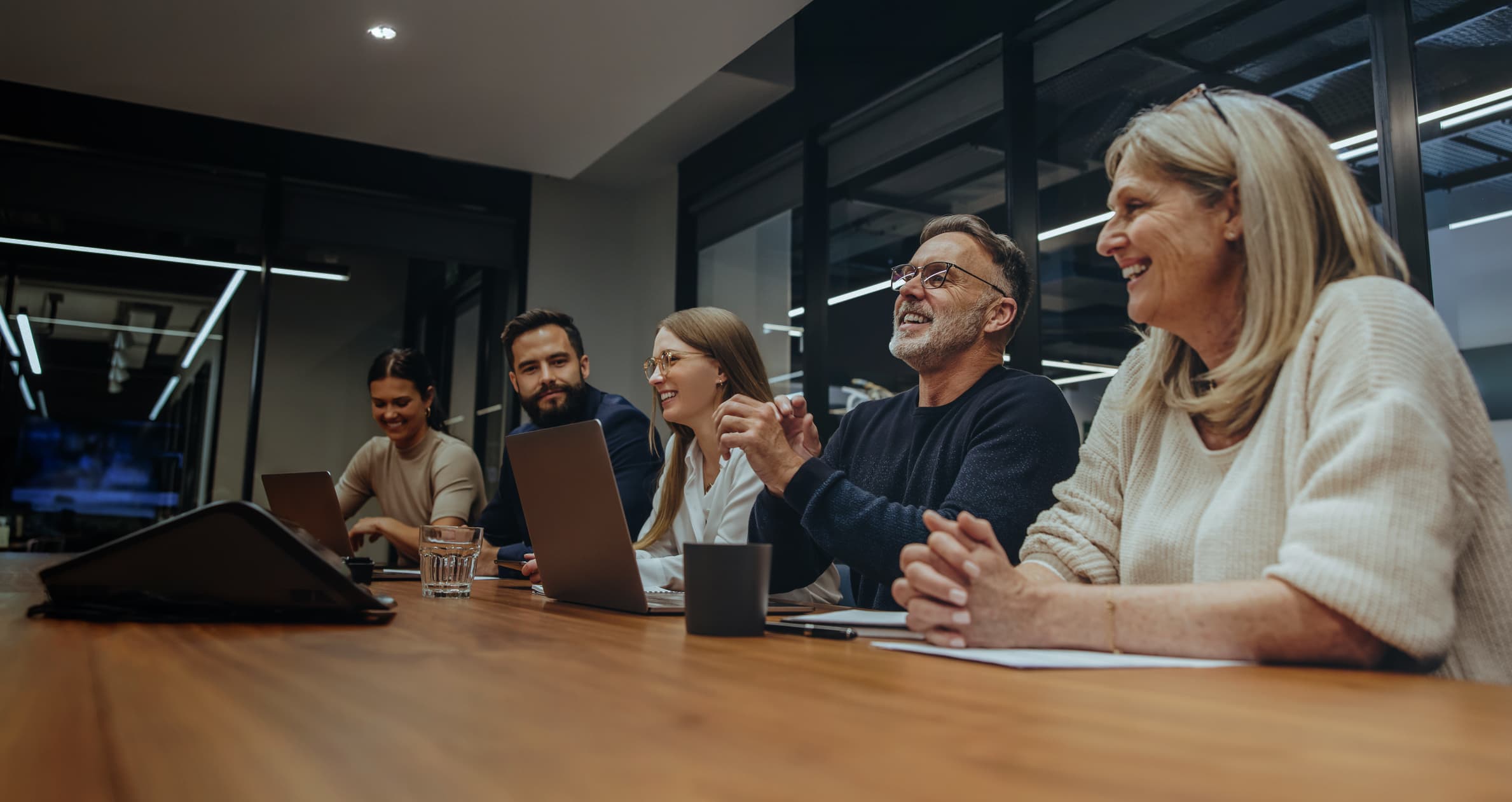 Ομάδα επαγγελματιών σε αίθουσα συσκέψεων συμμετέχει σε συνάντηση γύρω από τραπέζι, με laptops και σημειώσεις μπροστά τους.