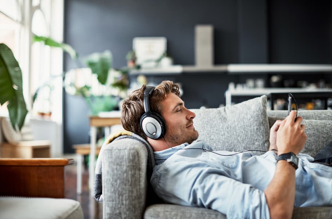 Άτομο ξαπλωμένο σε καναπέ φορά ακουστικά και κρατά κινητό τηλέφωνο, χαλαρώνοντας σε φωτεινό και άνετο καθιστικό.