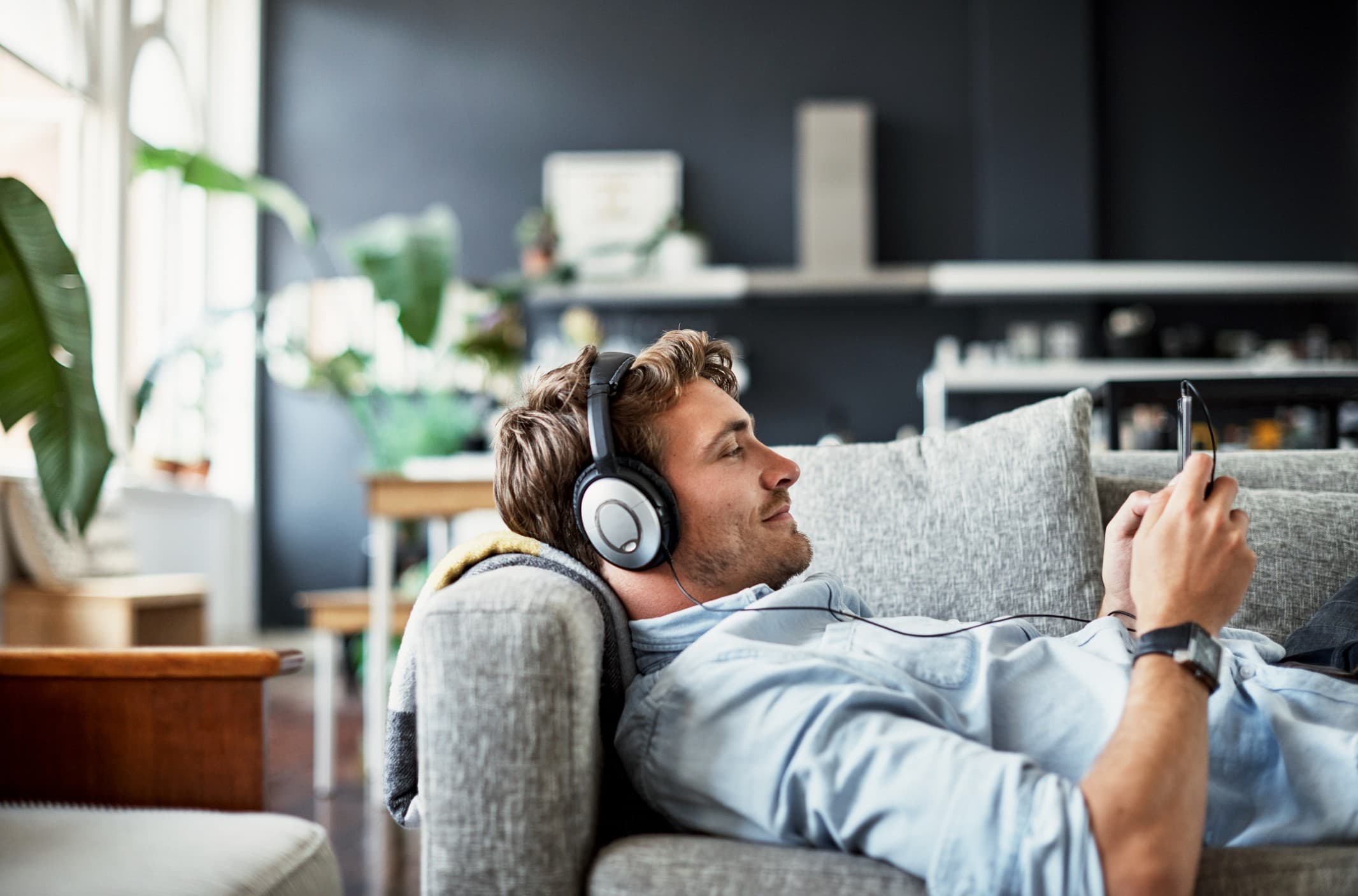 Άτομο ξαπλωμένο σε καναπέ φορά ακουστικά και κρατά κινητό τηλέφωνο, χαλαρώνοντας σε φωτεινό και άνετο καθιστικό.
